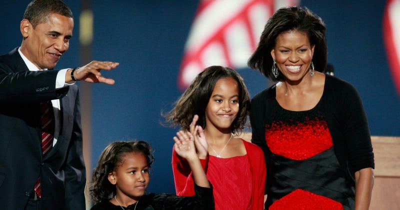 Barack Obama in a navy suit gesturing on stage alongside Sasha, Malia, and Michelle Obama — Malia in a red cardigan and Michelle in a black and red dress — at Barack Obama's 2008 election night victory celebration.