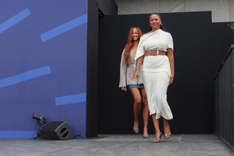 Malia and Sasha Obama walk on stage in front of a blue and black backdrop during what appears to be a fashion or entertainment event.