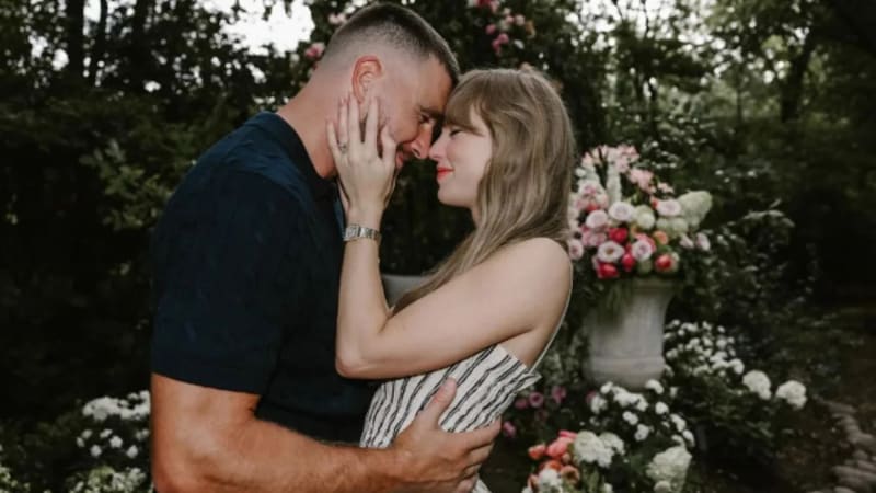 A man in a navy t-shirt and a woman in a striped strapless top face each other with foreheads nearly touching, embracing in a garden surrounded by pink and white floral arrangements.