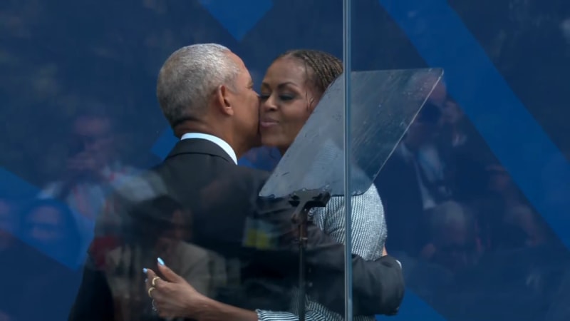 Barack Obama and Michelle Obama in formal attire greet each other warmly in front of a blue backdrop at what appears to be an official event.