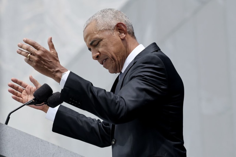 Barack Obama n a dark suit and white shirt speaks passionately at a microphone during an outdoor public address.