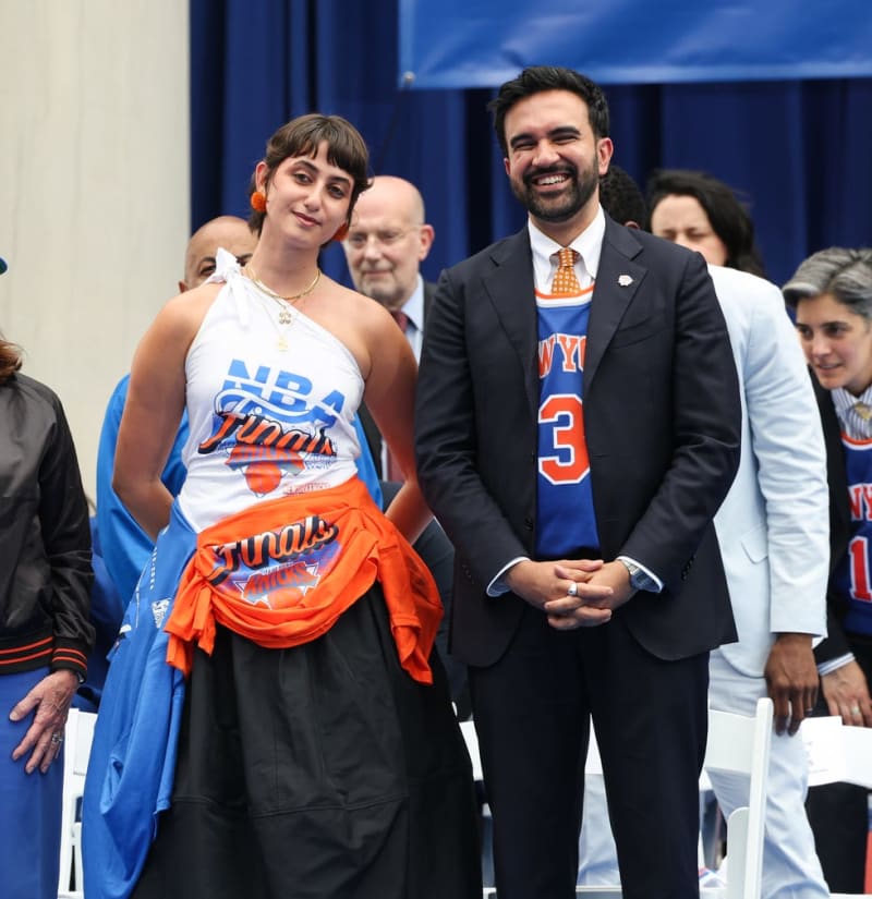 Rama Duwaji and Zohran Mamdani in New York Knicks apparel stand together at an indoor event, with spectators visible in the background.