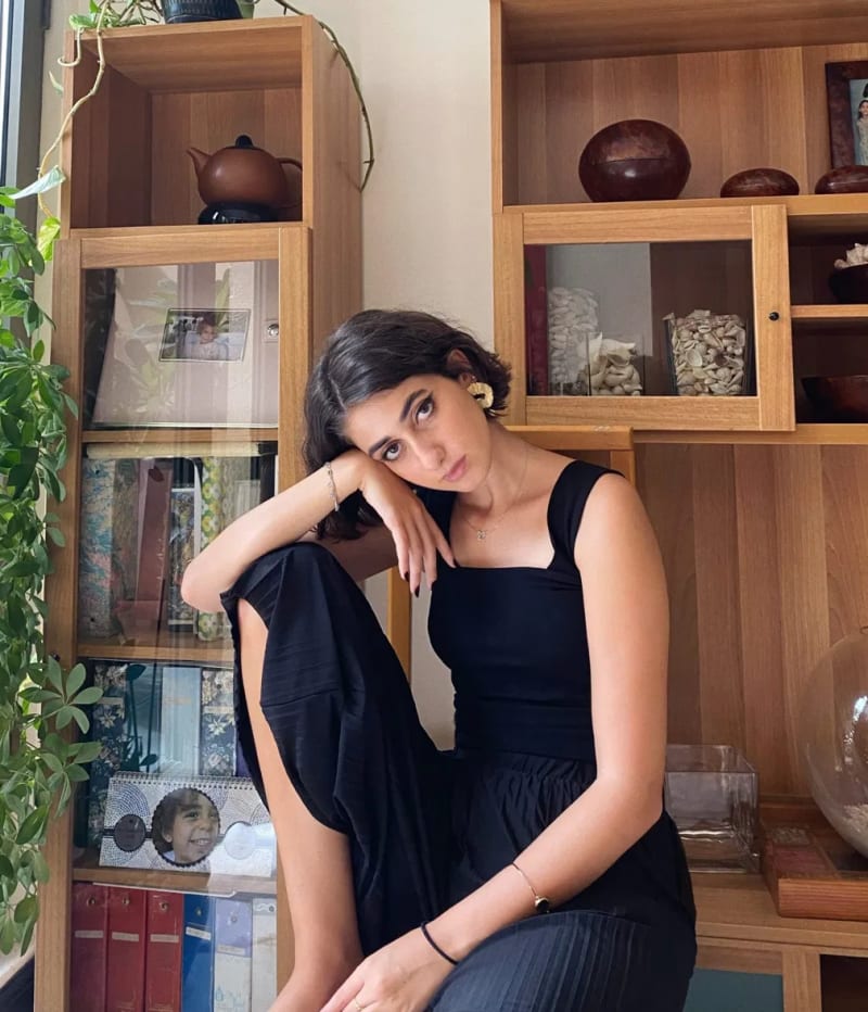 Rama Duwaji in a black sleeveless top with gold fan earrings and a delicate necklace, seated in front of wooden shelving units filled with books, ceramics, and plants.