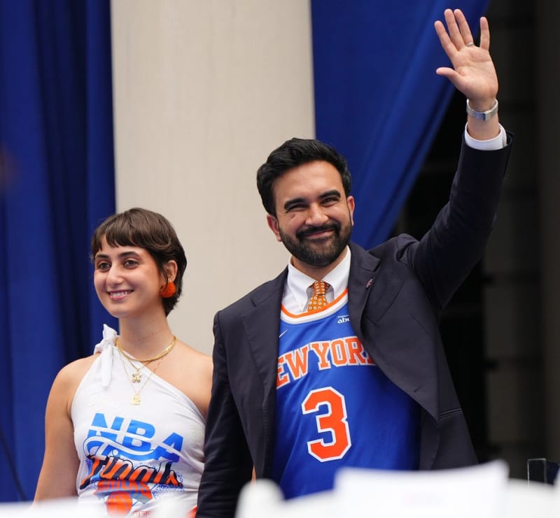 Rama Duwaji and Zohran Mamdani wearing New York Knicks apparel wave to a crowd at an event with blue curtains in the background.