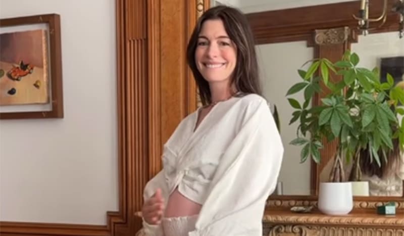 Anne Hathaway smiling and showing her pregnant belly in a white wrap top, standing indoors in front of a wooden mantel with a potted plant and framed artwork.