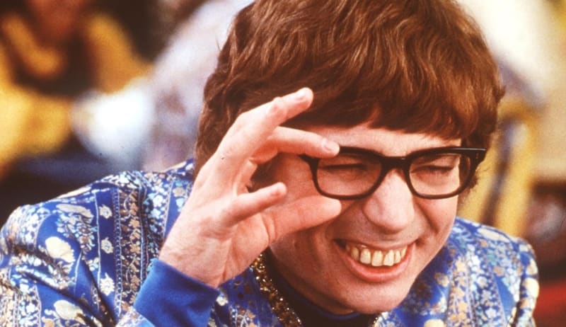 Mike Myers as Austin Powers, wearing glasses and a blue patterned jacket smiles while adjusting his eyeglasses with their hand.