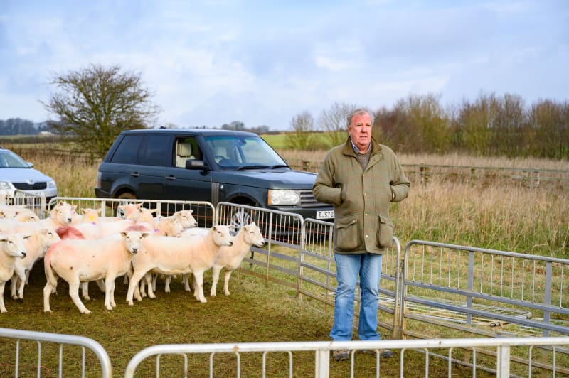 Jeremy Clarkson in a tan jacket stands among sheep pens at a rural farm event with vehicles and countryside in the background.