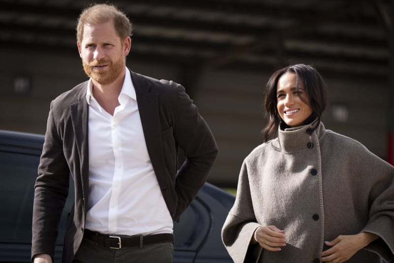 Prince Harry and Meghan Markle walking together outdoors, Harry in dark blazer and white shirt, Meghan in beige turtleneck sweater, both smiling.