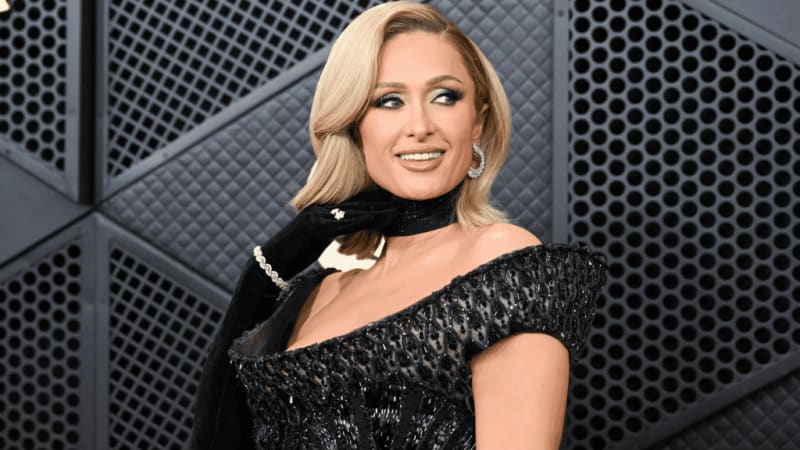 Paris Hilton with blonde hair wearing an off-shoulder black sequined dress and black choker against a geometric metallic background.