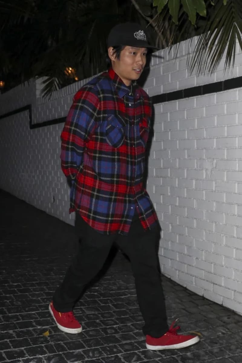 Pax Jolie-Pitt wearing a red and blue plaid shirt, black cap, and red sneakers stands against a white brick wall at night.
