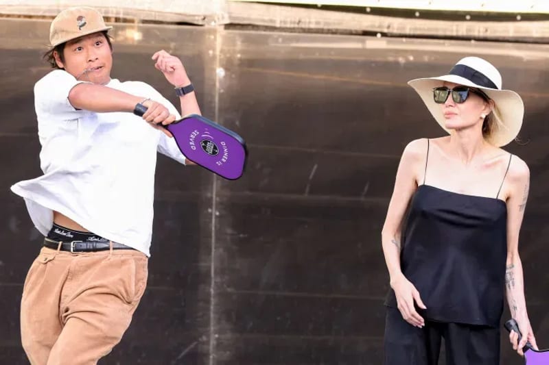 Pax Jolie-Pitt mid-swing with a paddle in a white t-shirt and cap, and Angelina Jolie wearing a wide-brim hat, sunglasses, and a black tank top, playing pickleball together.