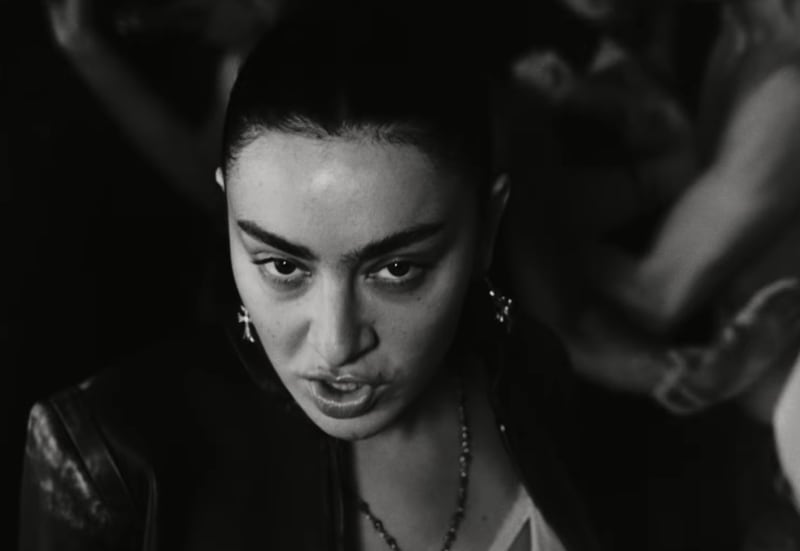 Charli XCX in dark clothing with pearl earrings gazes downward in this black and white portrait photograph.