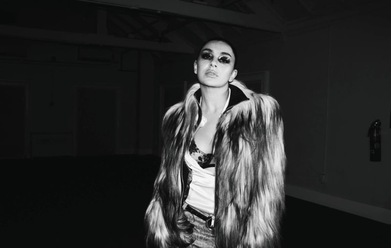 Charli XCX wearing a textured fur coat and bold eye makeup stands in a dimly lit interior space, looking upward.
