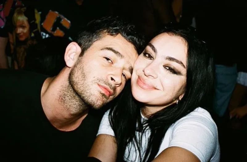 Matty Healy in a black top and Charli XCX in a white top with sunglasses pushed up on her head, posing close together for a selfie at a dark, crowded event.
