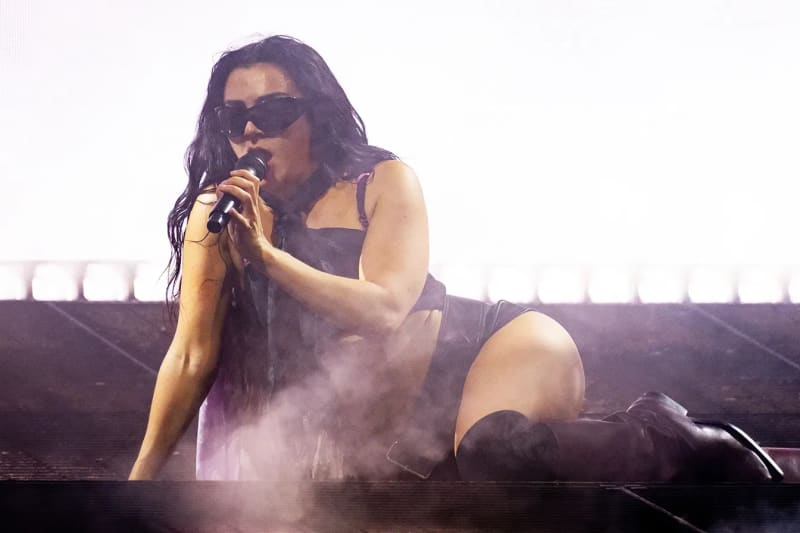 Charli XCX performing on stage in dark sunglasses and a black outfit with knee-high boots, kneeling while singing into a microphone with smoke effects around her at Glastonbury.