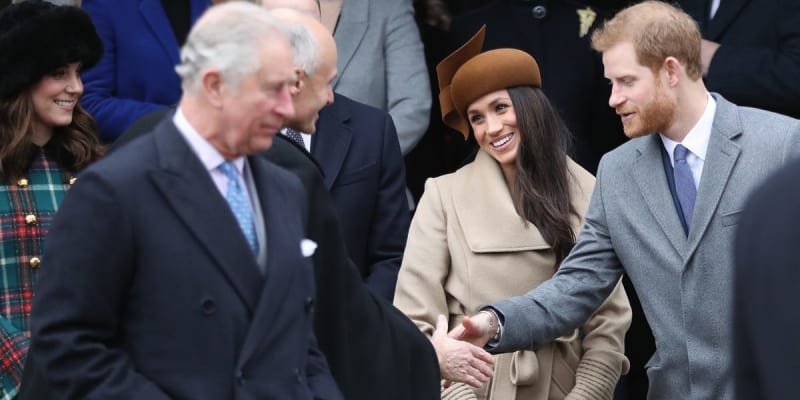 King Charles shaking hands with Prince Harry and Meghan Markle, who is wearing a camel coat and matching hat, with Kate Middleton visible in the background, gathered outdoors for a Christmas event.