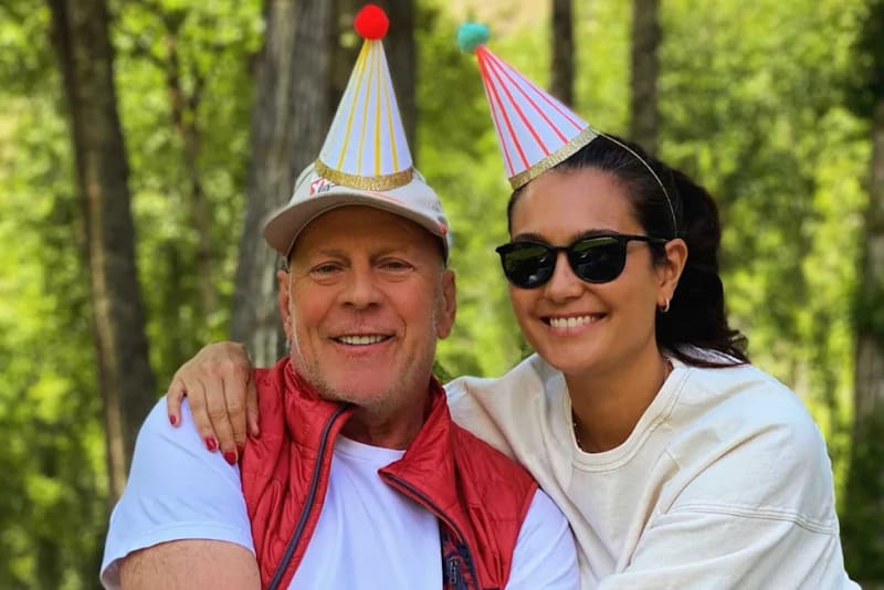 Bruce Willis and Emma Heming wearing colorful party hats smile together outdoors in front of green trees.