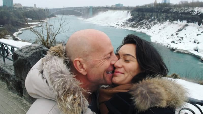 Bruce Willis and Emma Heming kissing at an overlook with Niagara Falls and snowy landscape visible in the background.