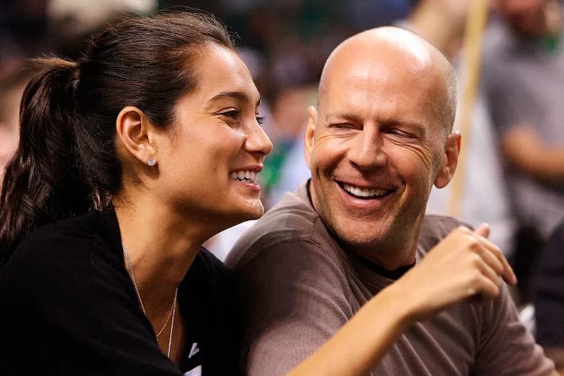 Emma Heming and Bruce Willis smiling closely together at what appears to be a sporting event, with a green and white crowd visible in the background.