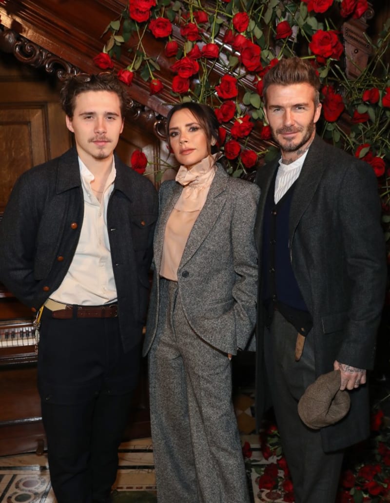 Brooklyn Beckham in a dark jacket and cream shirt, Victoria Beckham in a grey tweed suit, and David Beckham in a grey coat with navy vest stand together in front of a red rose display and wood-paneled staircase.