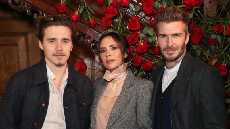 Brooklyn Beckham in a dark jacket and cream shirt, Victoria Beckham in a grey tweed suit, and David Beckham in a grey coat with navy vest stand together in front of a red rose display and wood-paneled staircase.