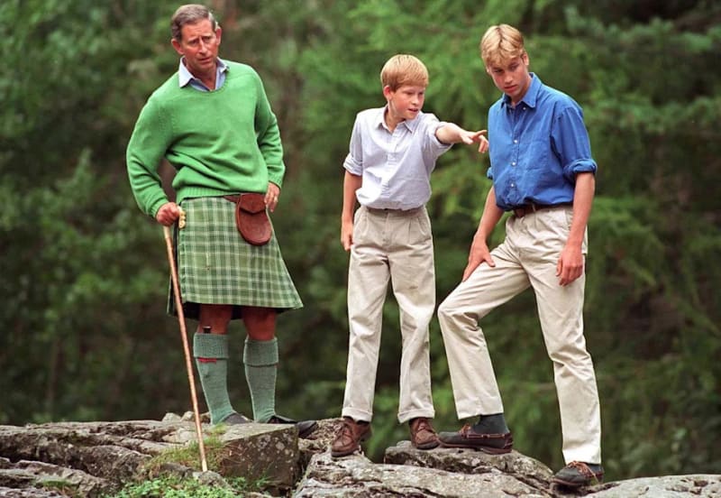 King Charles in a green sweater and tartan kilt with a walking stick stands with a young Prince Harry in a light blue shirt and young Prince William in a darker blue shirt, all standing on rocks in a wooded area.