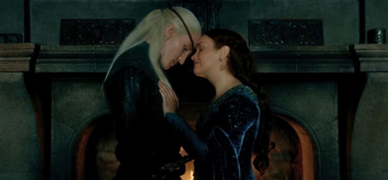 Two characters in a dimly lit stone room with a fireplace, one in black with long white hair leaning close to the other in a dark blue gown with dark hair.
