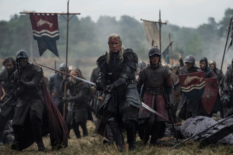 A warrior with long white hair in black armor leads armored soldiers across a battlefield with medieval banners and flags.