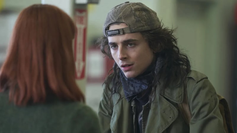 A young man with long dark hair in a backwards camo cap and olive jacket with a blue bandana around his neck, talking to a red-haired woman in a dimly lit indoor setting.