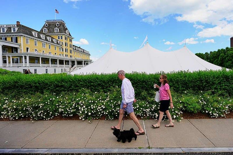 Two people walk a small black dog on a sidewalk past a large yellow hotel and white tent with flowering hedge behind.