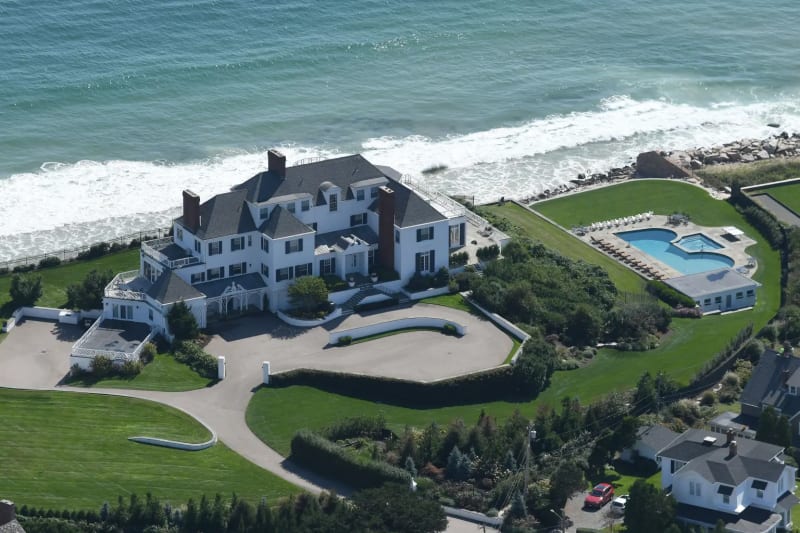 Aerial view of a large white Colonial-style mansion on a seafront estate with a pool, manicured lawns, and a private driveway, with the ocean directly behind.