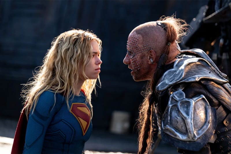A woman in a Superman costume faces a man with facial tattoos and armor in an intense confrontation scene.