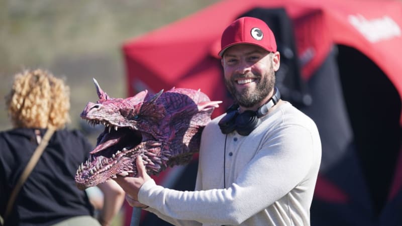 A bearded man in a red cap holds a purple dragon figurine while smiling at an outdoor event with a red vehicle behind him.