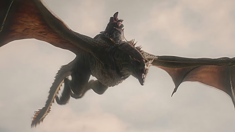 A dragon rider in black armour soars through an overcast sky on a large dark dragon with wide-spanning wings.
