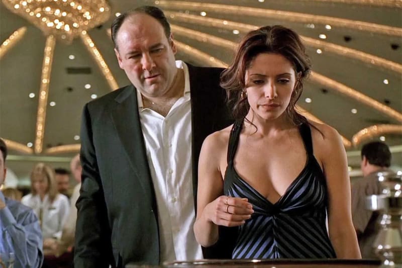 A man in a dark suit jacket and open-collar shirt stands close behind a woman in a striped halterneck dress at a casino table, with chandeliers and other patrons visible in the background.