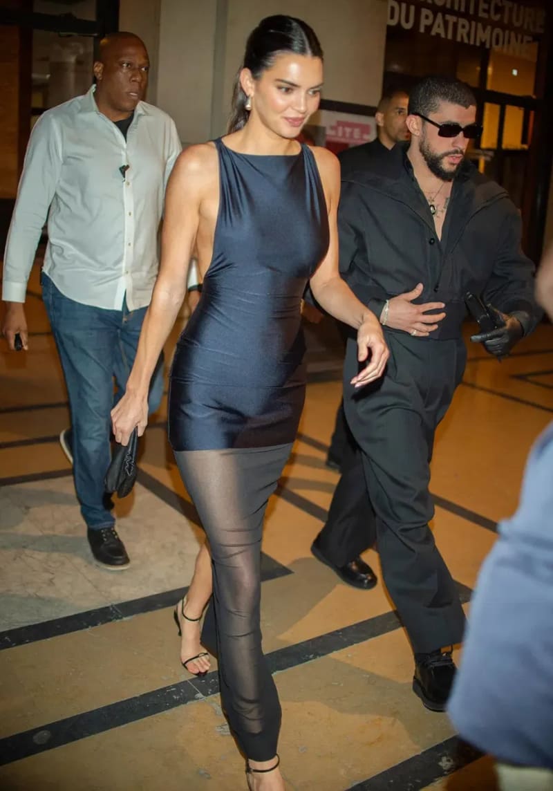 A woman in a navy bodycon dress and sheer leggings walks alongside a man in an all-black outfit and sunglasses, with security personnel visible behind them.
