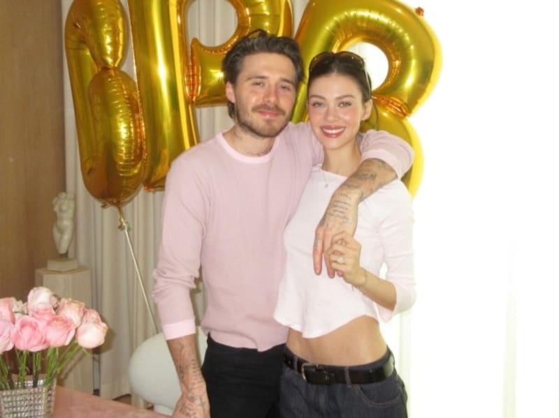 A couple poses together smiling in front of gold number balloons, with pink flowers visible on the left side.