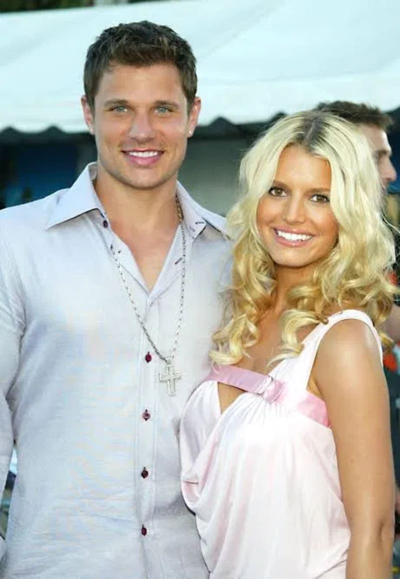A man in a light grey shirt with a cross necklace stands next to a woman with long blonde curly hair in a pink dress at an outdoor event.