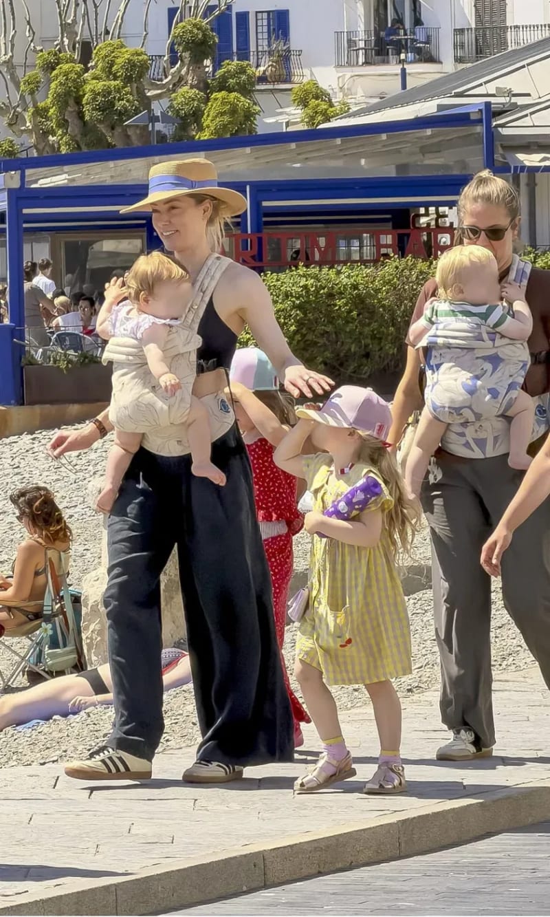 A woman in a straw hat, black top and wide-leg trousers carrying a baby in a front carrier, with a young girl in a yellow dress beside her and another woman carrying a baby behind them, on a sunny seafront promenade.