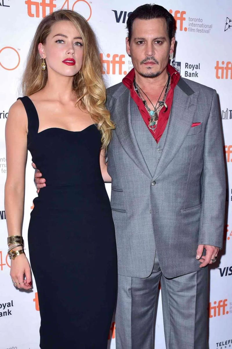 A woman in a black fitted dress with gold bracelets and a man in a grey suit with a red shirt and layered necklaces, posing together on a red carpet at the Toronto International Film Festival.