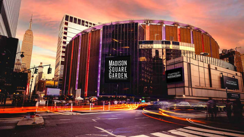 Exterior of Madison Square Garden at dusk, with light trails from passing traffic in the foreground and the Empire State Building visible in the background.