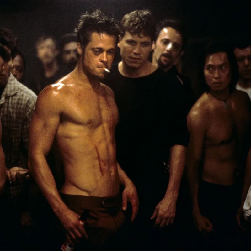 A shirtless man with a cigarette in his mouth, visible cuts on his torso, stands in front of a crowd of men in a dimly lit basement setting.