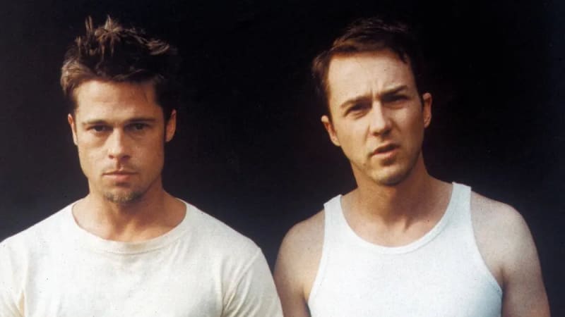 Two men in white tops stand side by side against a dark background — one in a crew-neck t-shirt and the other in a sleeveless vest