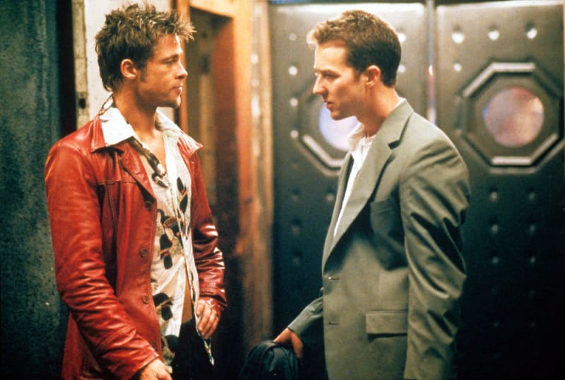 A man in a red leather jacket and open patterned shirt faces another man in a grey suit, both standing in a dimly lit industrial corridor.