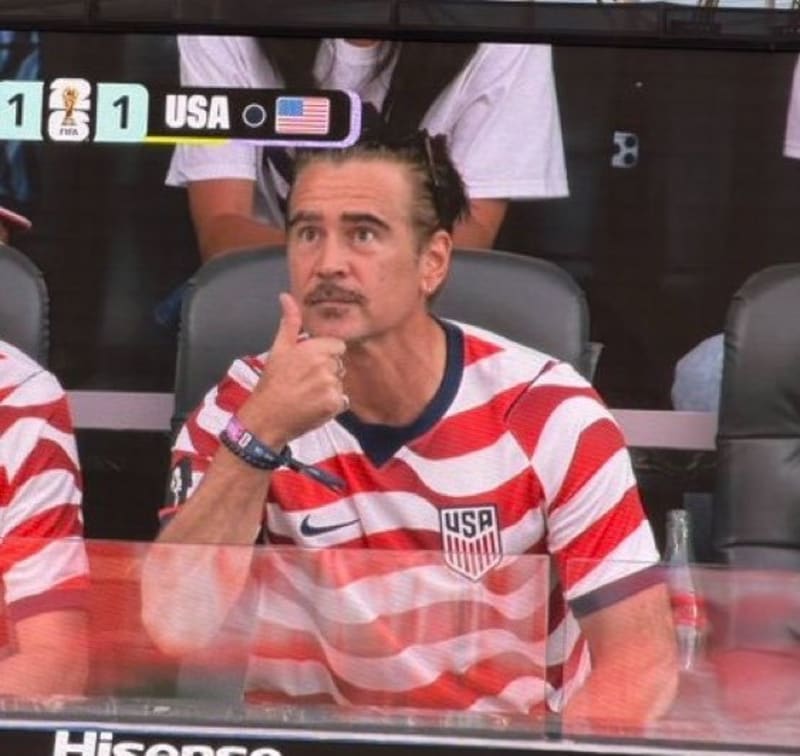 Man in a USA soccer jersey giving a thumbs up in stadium seating, FIFA World Cup scoreboard visible behind him.