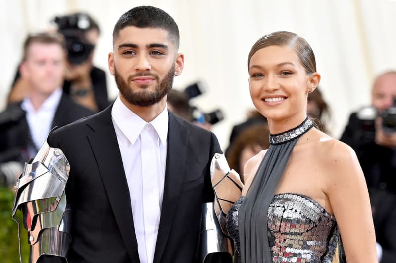 A man in a black suit with silver armour panels at the wrists stands beside a woman in a silver sequined halter gown, both on a red carpet.