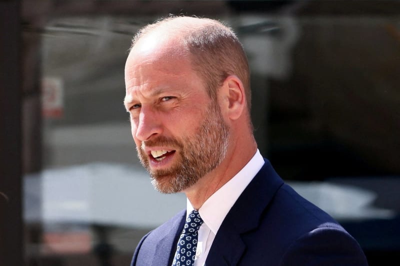A man with receding hair and a beard wearing a dark blue suit and patterned tie smiles while looking to the side.