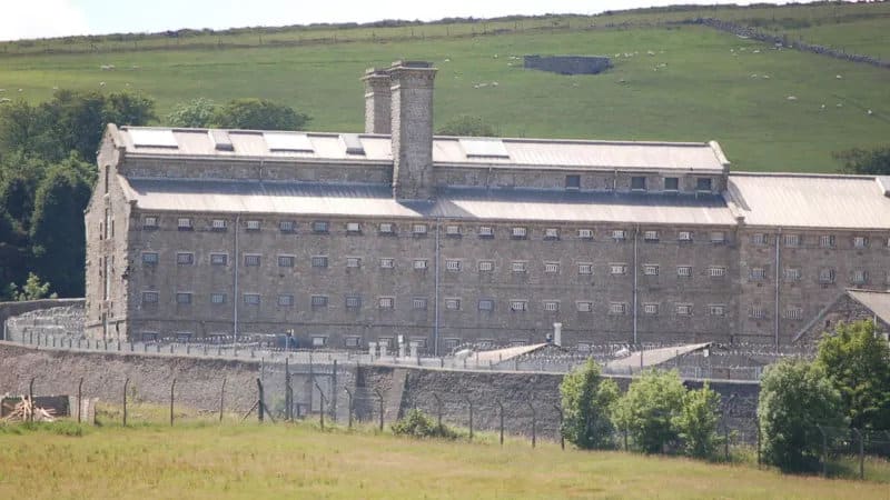A large Victorian granite prison building behind perimeter walls and razor wire fencing, set against green moorland hillside.