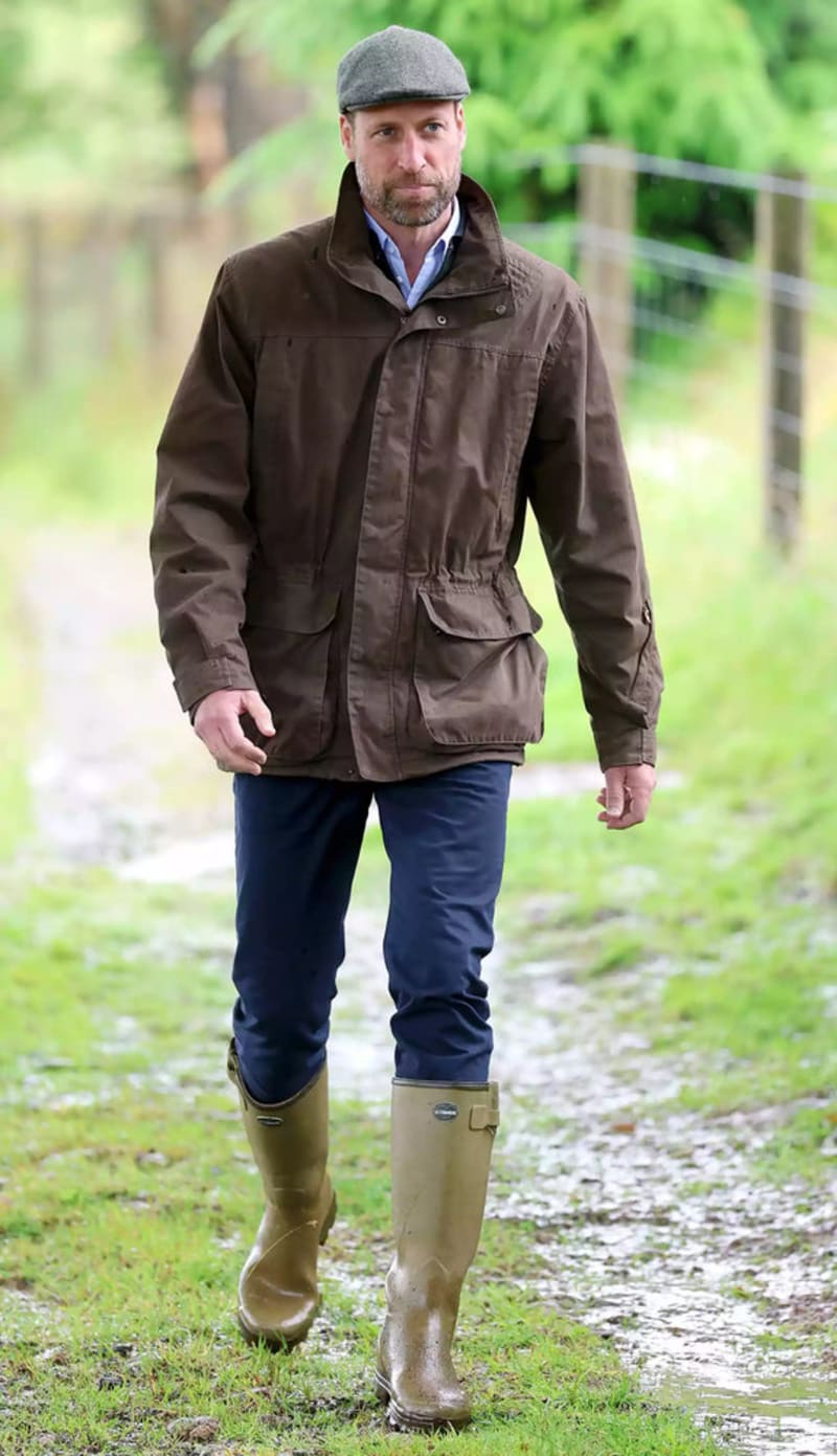 A bearded man in a brown wax jacket, flat cap, navy trousers and green wellington boots walks along a muddy rural path.