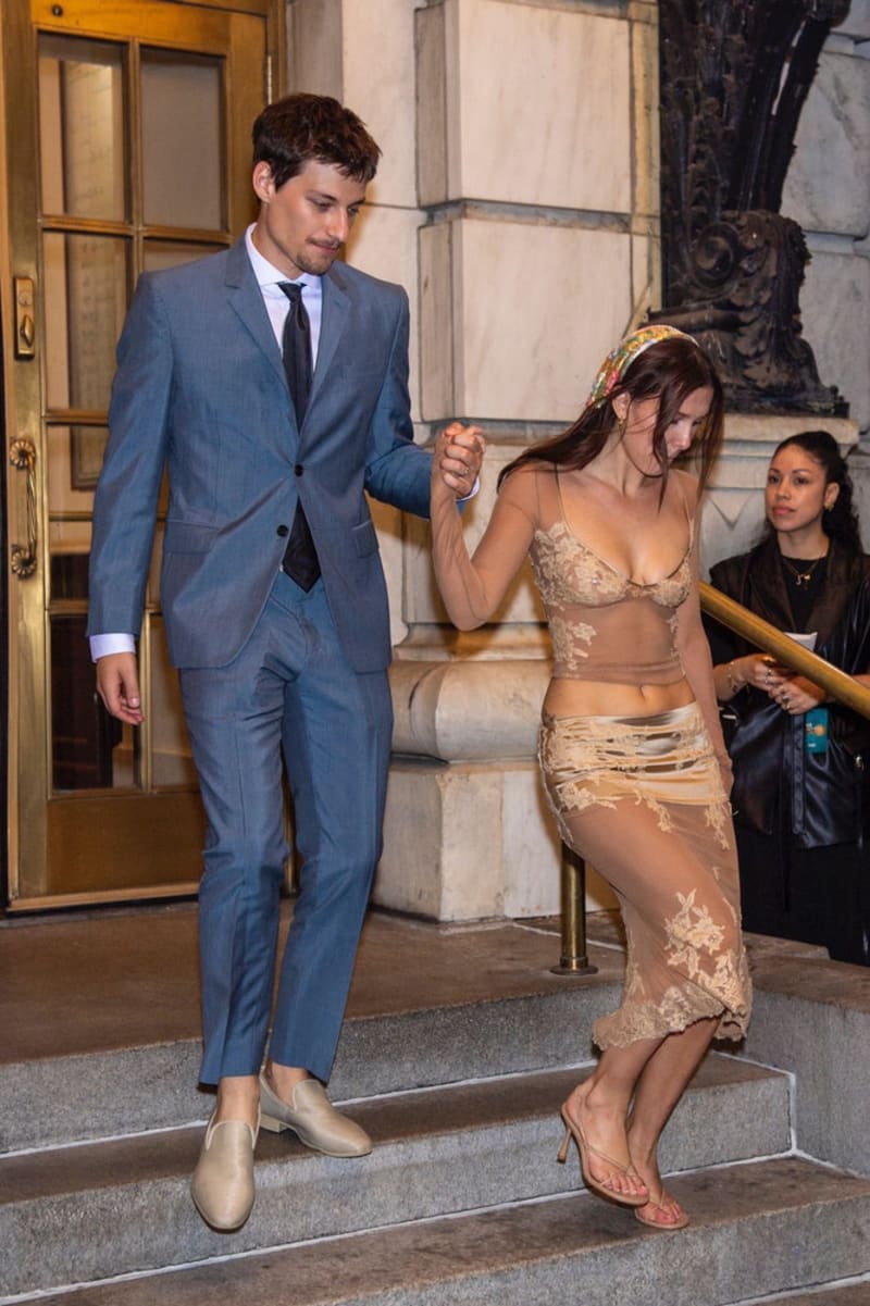A man in a blue suit and dark tie holds the hand of a woman in a sheer nude lace top and skirt with a beaded headband as they descend stone steps outside a grand building.